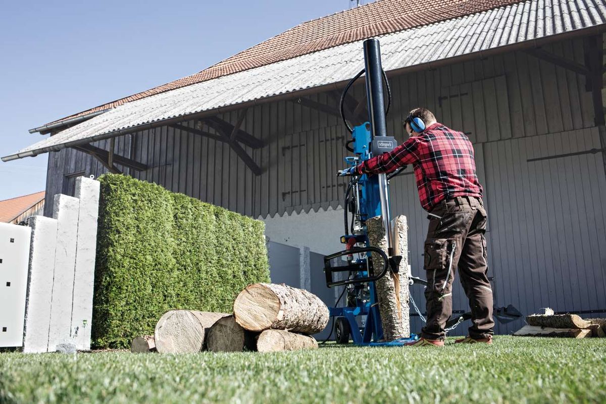 Ein Mann bedient einen Meterholzspalter HL3000GM von Scheppach. Er trägt Schutzausrüstung und schiebt einen Holzstamm in die Maschine. Im Hintergrund befindet sich ein Gebäude mit einer Hecke. Der Holzspalter ist blau und weiß.