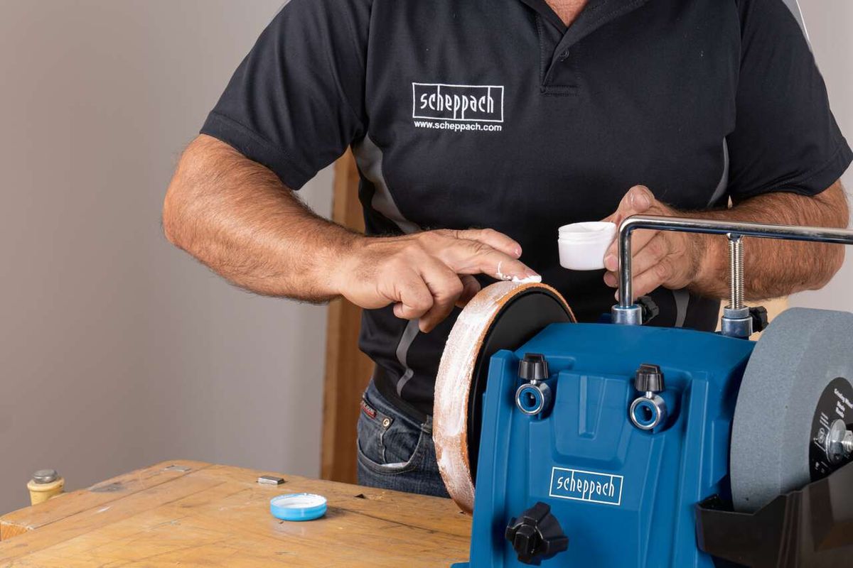A man operates a Scheppach wet bench grinder. He holds a grinding wheel and a pot of water in his hands. The machine has a blue and white grinding wheel.
