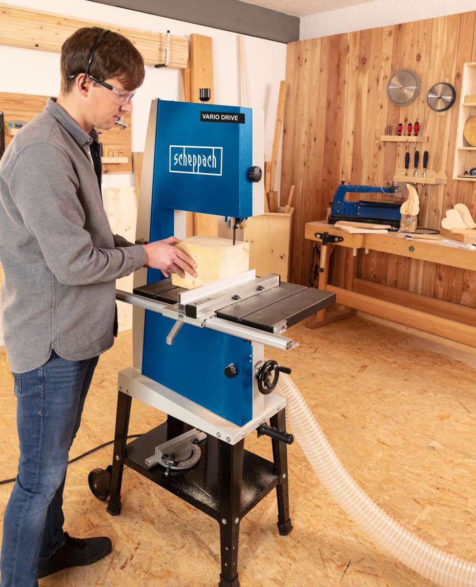 Ein Mann bedient eine Bandsäge von Scheppach in einer Werkstatt. Die Maschine hat die Bezeichnung 'Vario Drive'.
