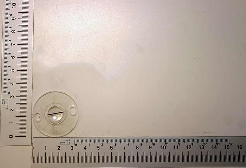Ein Lineal mit Millimeter- und Zentimeter-Einteilung. Im Hintergrund ist ein runder Gegenstand mit einer Linse zu sehen.