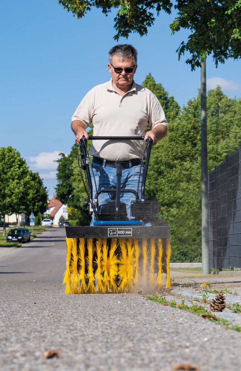 Ein Mann bedient eine Kehrmaschine SC2400p von Scheppach auf einem Gehweg. Die Maschine hat eine Arbeitsbreite von 600 mm.