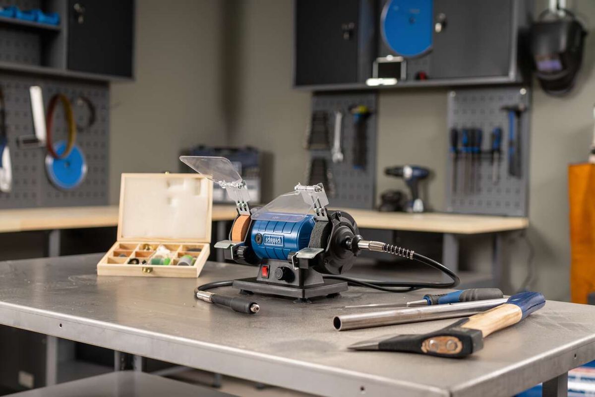 Doppelschleifer HG35 von Scheppach mit Zubehör auf einer Werkbank. Im Vordergrund liegen verschiedene Schleifwerkzeuge und eine Holzkiste mit Schleifscheiben.