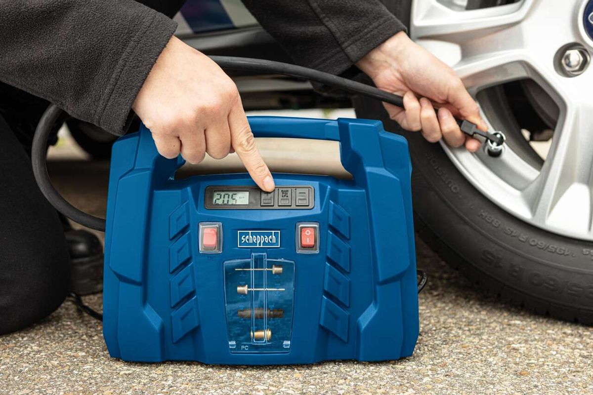 Ein Scheppach Kompressor mit LCD-Display und Druckanzeige. Eine Hand hält den Kompressor, während die andere ein Werkzeug hält. Der Kompressor ist mit einem Auto verbunden.