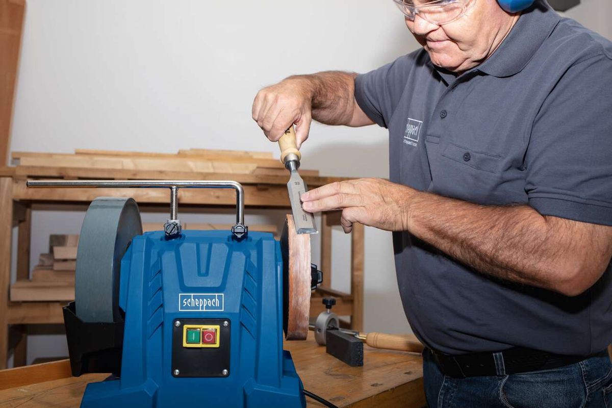 A man is sharpening a knife using the Scheppach Wet Bench Grinder Tiger 7000S. He holds the knife against the rotating grinding wheel, carefully guiding it along the blade.