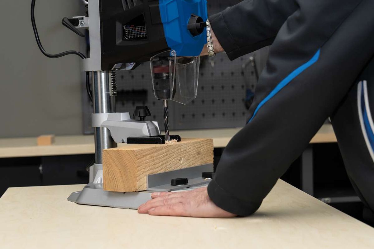 Tischbohrmaschine DP60 Scheppach im Einsatz. Ein Holzblock wird auf der Arbeitsplatte fixiert und mit einem Bohrer bearbeitet.