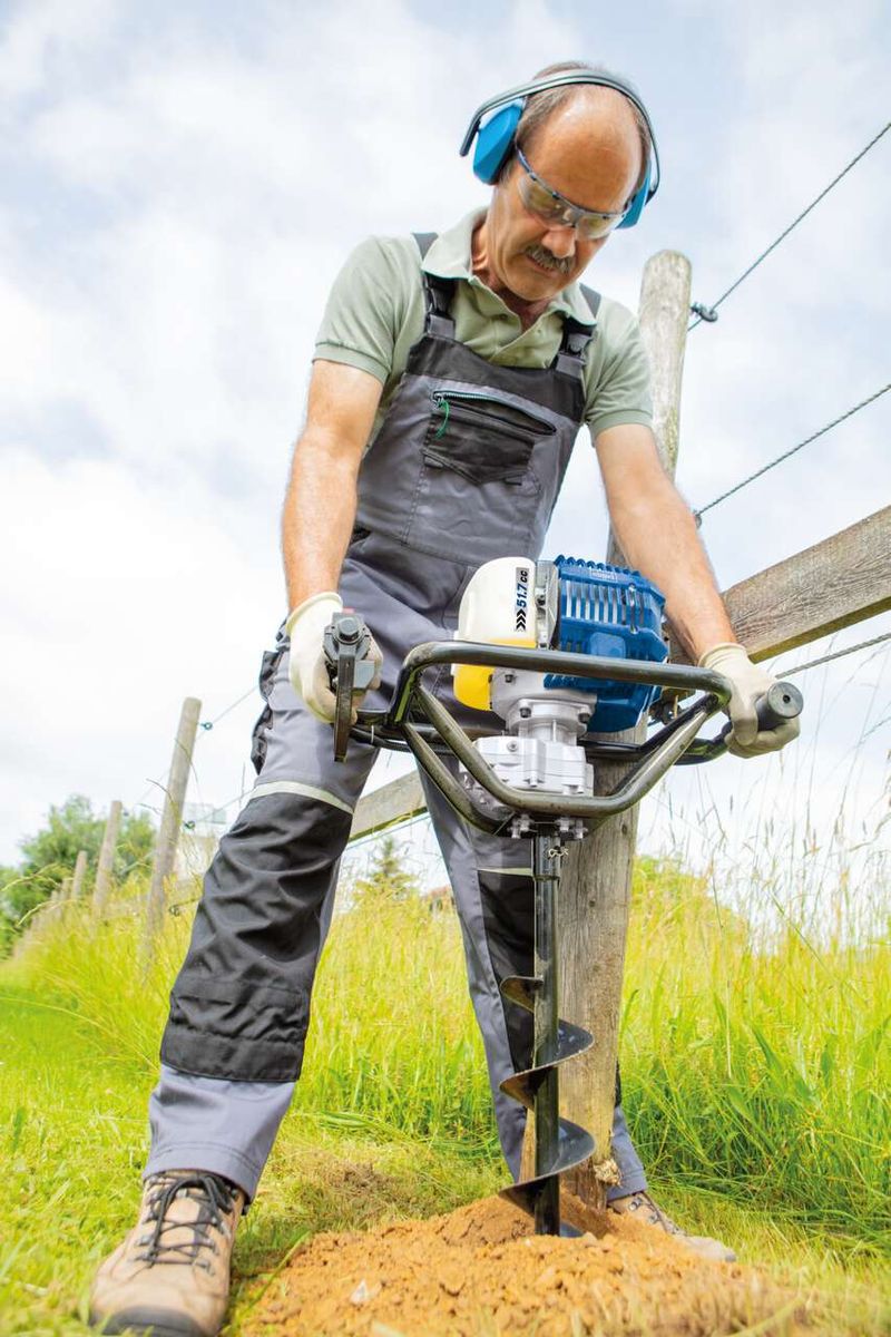 Ein Arbeiter verwendet den Benzin Erdbohrer EB1700 von Scheppach zum Graben eines Lochs. Der Arbeiter trägt Schutzausrüstung und hält den Bohrer mit beiden Händen fest. Der Bohrer hat eine Bohrlänge von 800 mm und ist Teil eines Bohrersets.