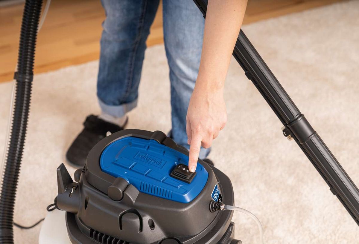 Ein Scheppach-Sprüh- und Waschsauger wird eingeschaltet. Die Person drückt den schwarzen Knopf auf dem blauen Deckel des Geräts. Der Boden ist mit einem hellen Teppich bedeckt.