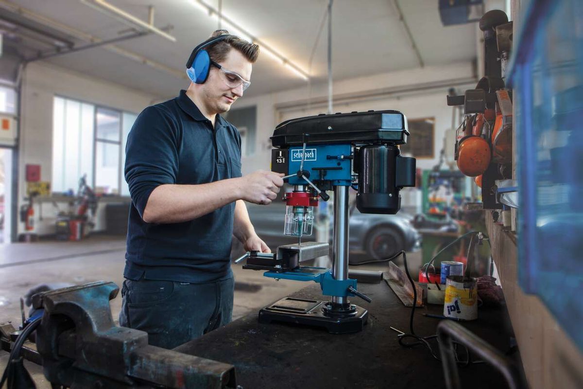 A man operates a Scheppach bench drill DP16VLS in a workshop. He is wearing safety goggles and ear protection. The machine features a laser for precise drilling.