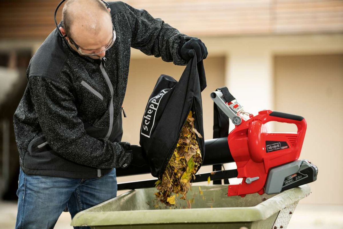 Ein Mann leert einen mit Laub gefüllten Beutel in einen Scheppach Laubsauger und -bläser. Der Laubsauger und -bläser BBV500-40Li von Scheppach ist auf einem Wagen montiert.