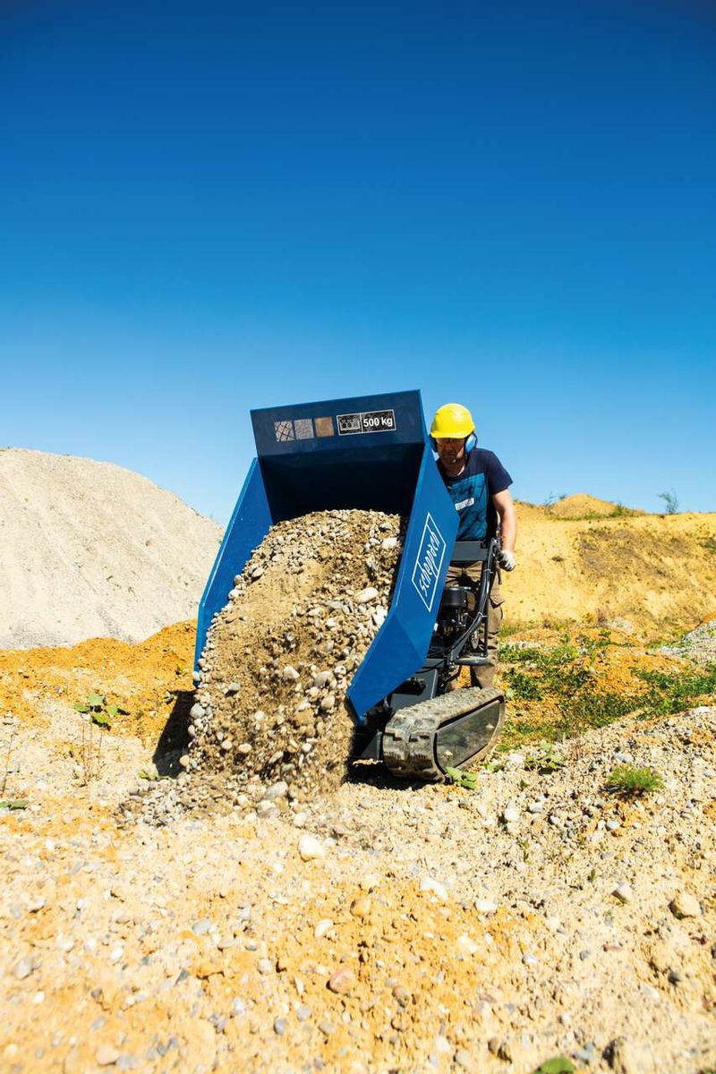 Kettendumper DP5000 Scheppach mit gelbem Helm tragender Person. Der Dumper ist mit Steinen beladen und fährt auf einem Kiesweg.