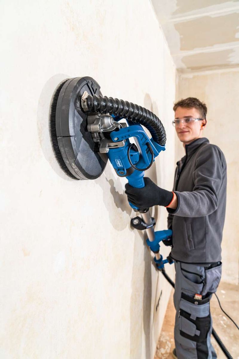 Ein Mann verwendet den Wand- und Deckenschleifer DS920X von Scheppach an einer Wand. Der Schleifer hat einen Durchmesser von 225 mm und ist mit einem 4 m langen Schlauch verbunden.