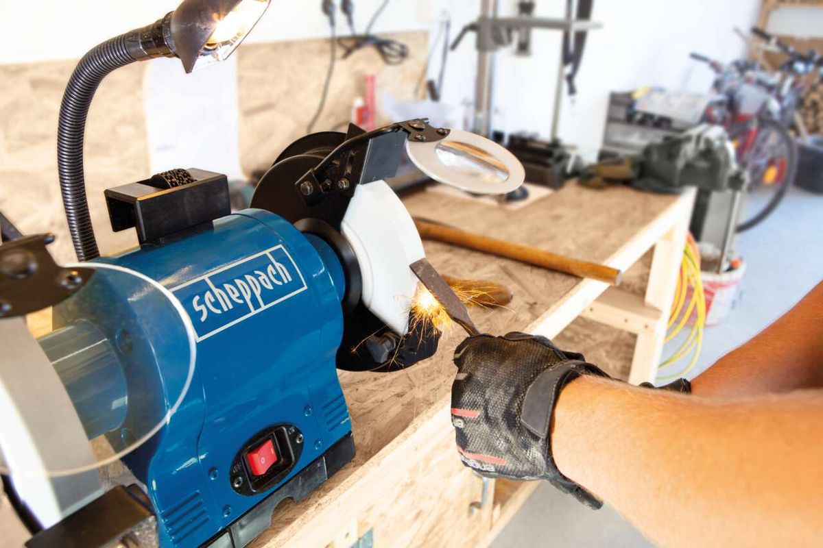 Scheppach Doppelschleifer BG200AL im Einsatz. Ein Handschuh hält ein Metallstück gegen die Schleifscheibe. Im Hintergrund sind Werkzeuge und Fahrräder zu sehen.