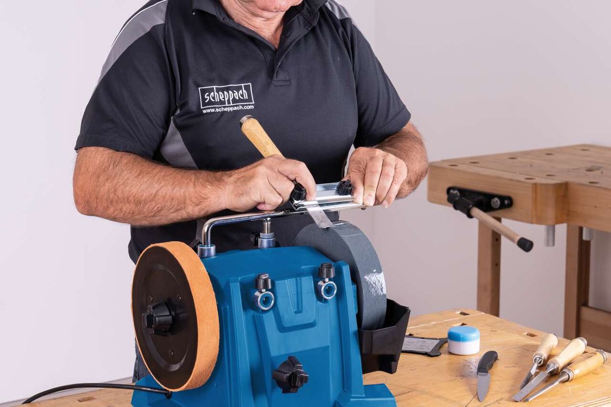 Ein Mann bedient eine Scheppach Nass-Schleifmaschine. Er hält einen Holzgriff in der Hand und justiert die Maschine. Im Hintergrund ist eine Werkbank mit verschiedenen Werkzeugen zu sehen.