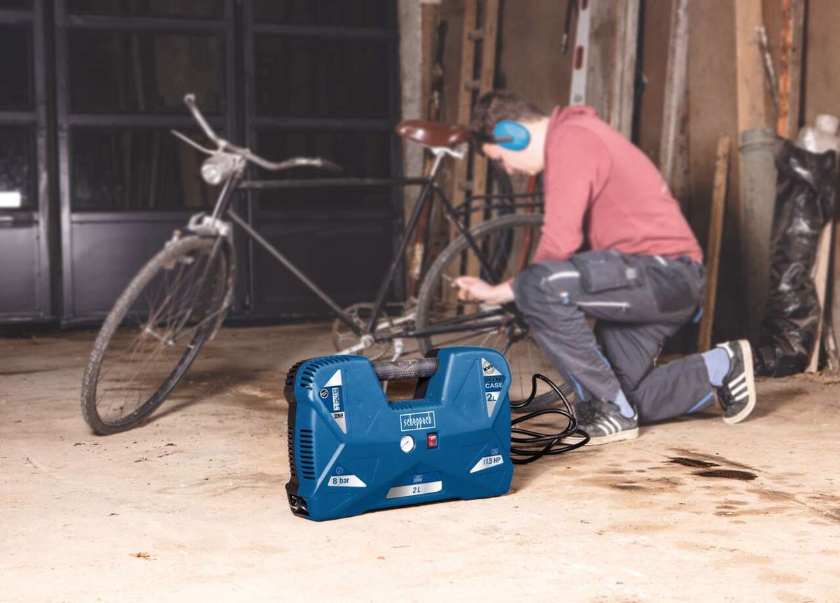 Ein Mann kniet auf dem Boden und repariert ein Fahrrad. Neben ihm steht ein Scheppach Kompressor. Im Hintergrund sind Werkzeuge und Materialien zu sehen.