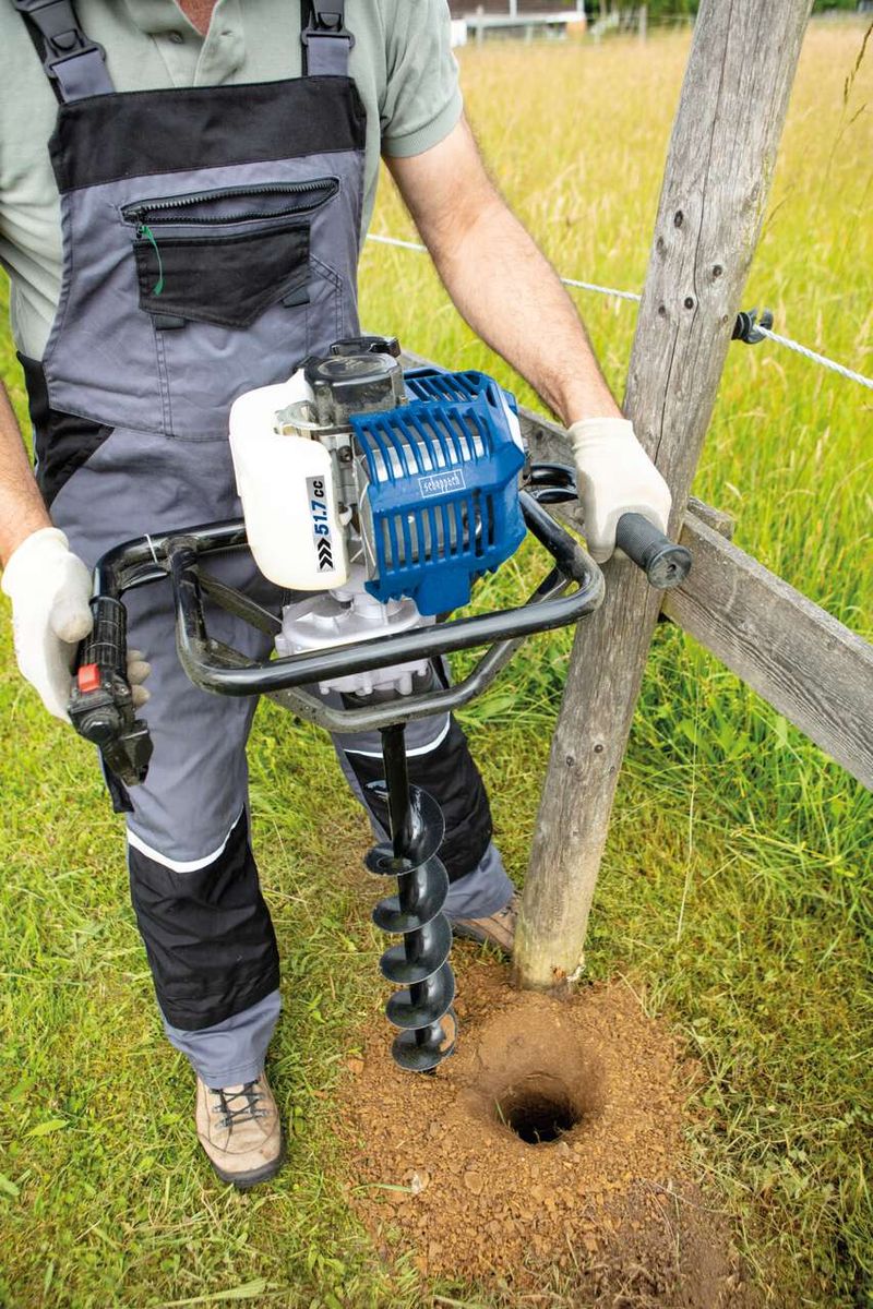 Ein Arbeiter verwendet einen Benzin-Erdbohrer von Scheppach, um ein Loch in den Boden zu graben. Der Erdbohrer hat eine Bohrkrone und wird von einem Motor angetrieben. Der Arbeiter trägt Schutzausrüstung, darunter Handschuhe und robuste Kleidung.