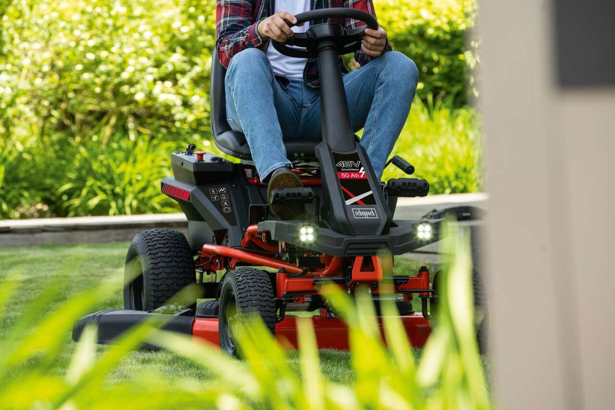 Akku-Aufsitzrasenmäher MR-760E von Scheppach im Einsatz. Person sitzt auf dem Mähgerät und steuert es auf einem Rasen. Das Gerät hat LED-Licht und einen Tempomat.