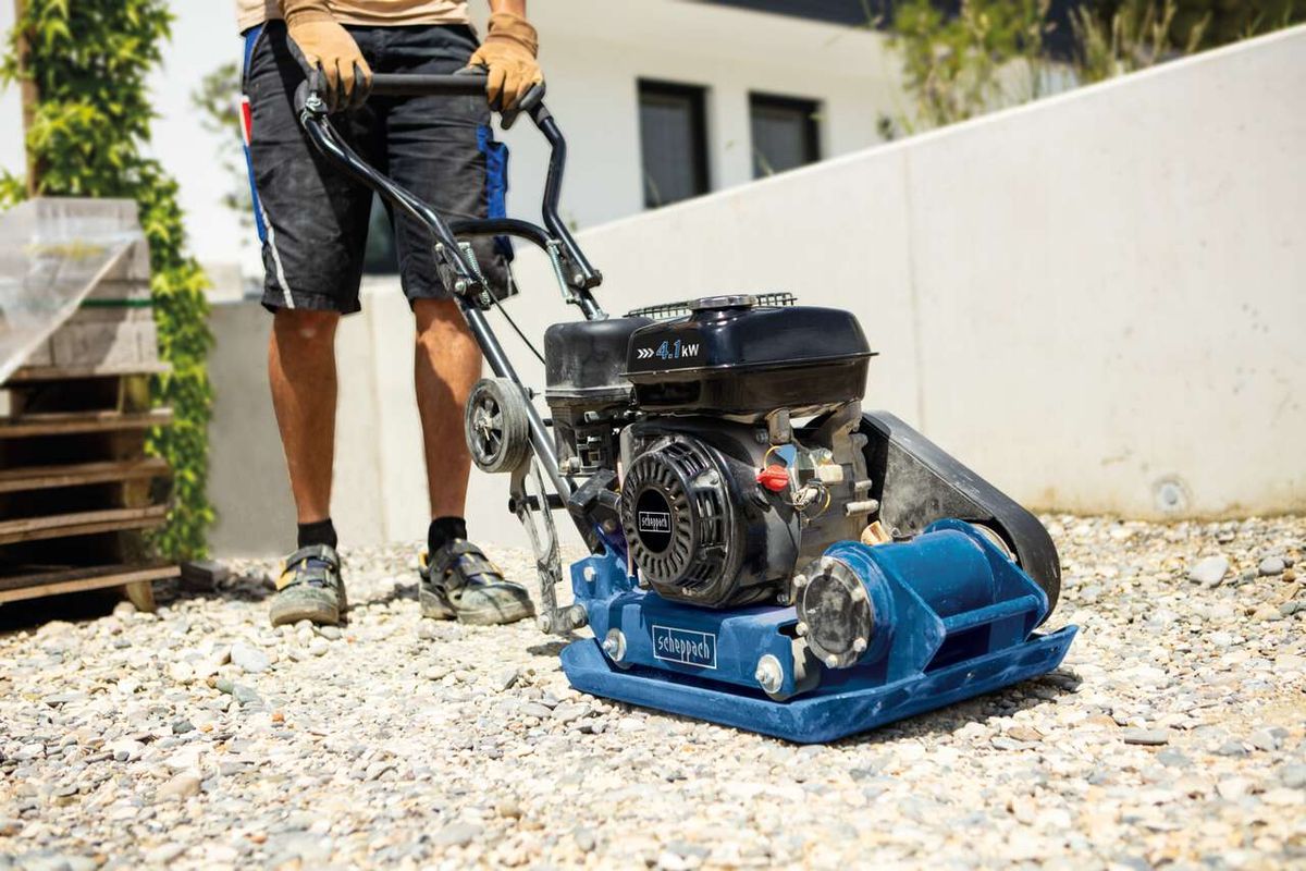 Ein Mann bedient eine Scheppach Rüttelplatte HP1300S auf einem Kiesboden. Die Maschine hat einen schwarzen Motor und einen blauen Rahmen. Der Name 'Scheppach' ist auf dem Gehäuse sichtbar.