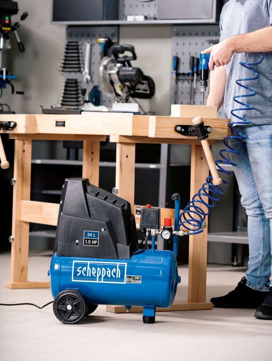 Scheppach HC300 compressor with accessories. A man is using an air drill at a workbench. Various tools and equipment are visible in the background.