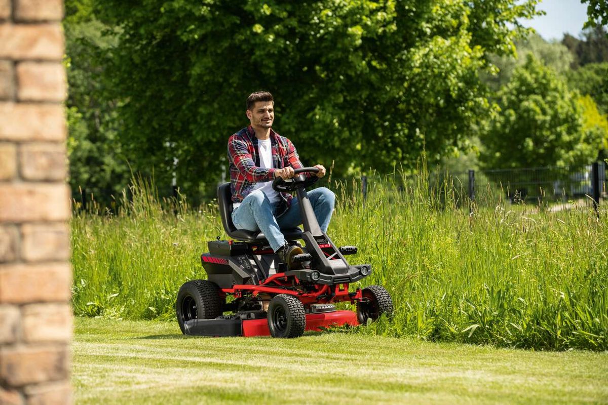 Ein Mann sitzt auf einem Akku-Aufsitzrasenmäher MR-760E von Scheppach und mäht einen Rasen. Der Mäher hat eine Schnittbreite von 76 cm und ist mit LED-Licht und Tempomat ausgestattet.