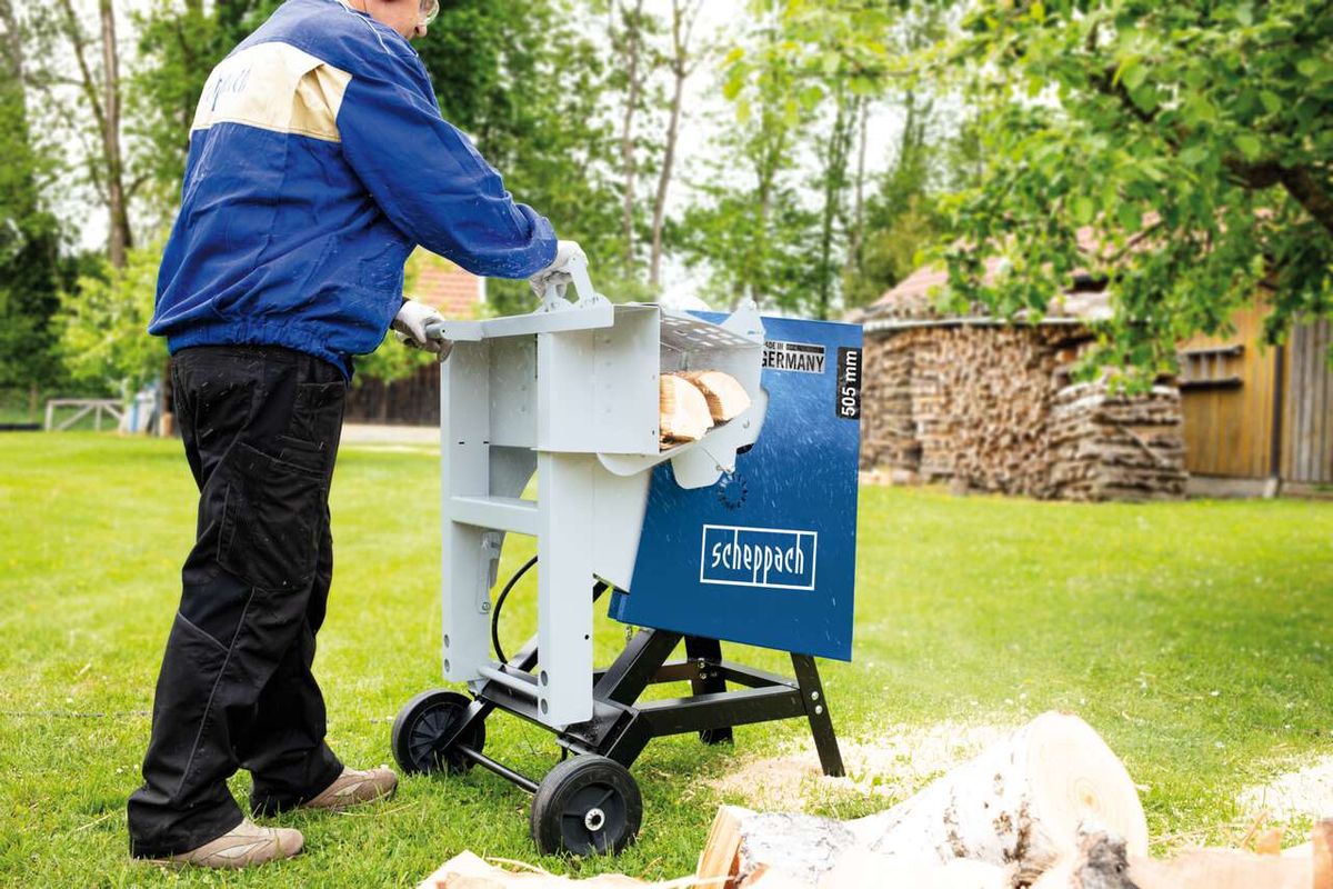 Ein Mann bedient eine Wippsäge HS520 von Scheppach im Freien. Die Säge ist auf einem grünen Rasen platziert, mit Holzstämmen daneben. Im Hintergrund sind Bäume und ein Holzstapel zu sehen.