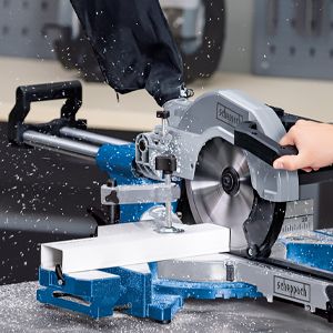 A Scheppach tool cutting wood. A person holds the wood steady while the saw cuts it. The saw is mounted on a table and is cutting the wood in a workshop setting.