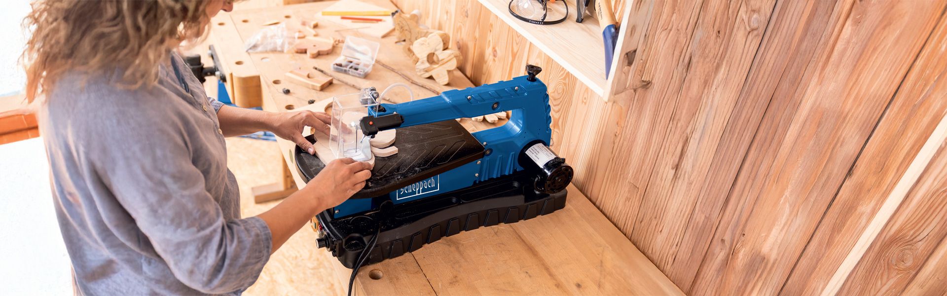 A person is operating a Scheppach DecoFlex saw in a workshop. The saw is placed on a wooden table, with various tools and materials visible in the background.