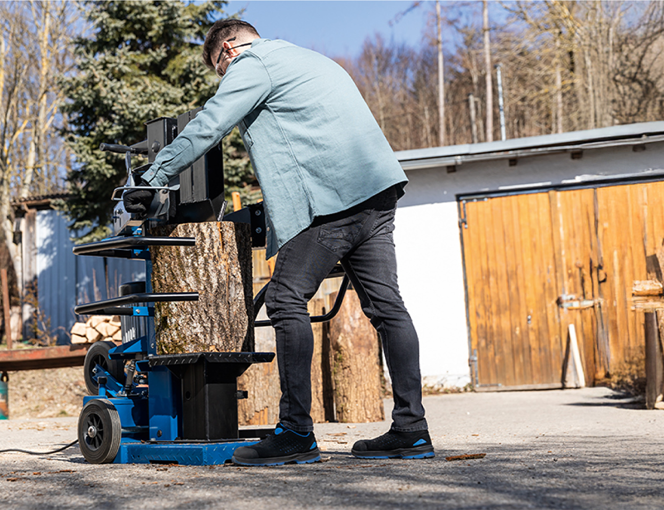 Ein Mann bedient einen mobilen Holzspalter von Scheppach im Freien. Der Holzspalter hat einen großen Holzklotz darin. Die Szene spielt vor einem Schuppen mit Holzstapel im Hintergrund.