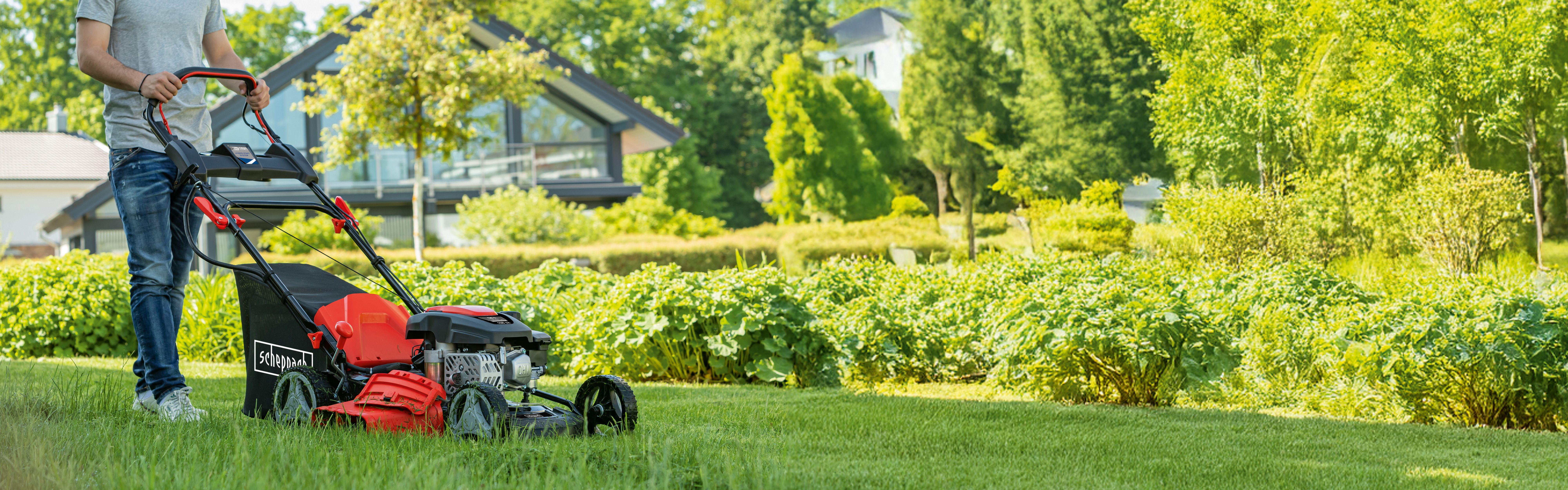 Mann mit Scheppach-Rasenmäher auf grünem Rasen vor Haus mit Garten
