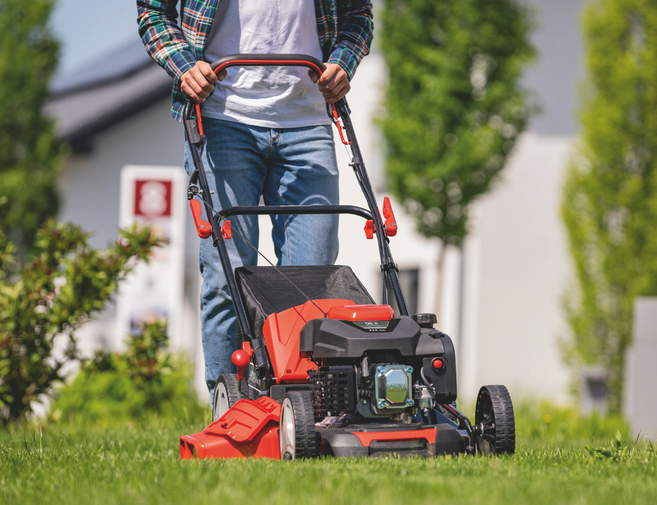 Ein Mann schiebt einen Rasenmäher von Scheppach über eine Wiese. Der Rasenmäher hat einen schwarzen Auffangbehälter und einen roten Motor.