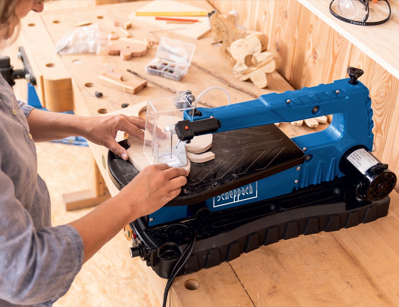 A man operates a Scheppach DecoFlex machine in a workshop. The workbench is covered with woodwork and tools.