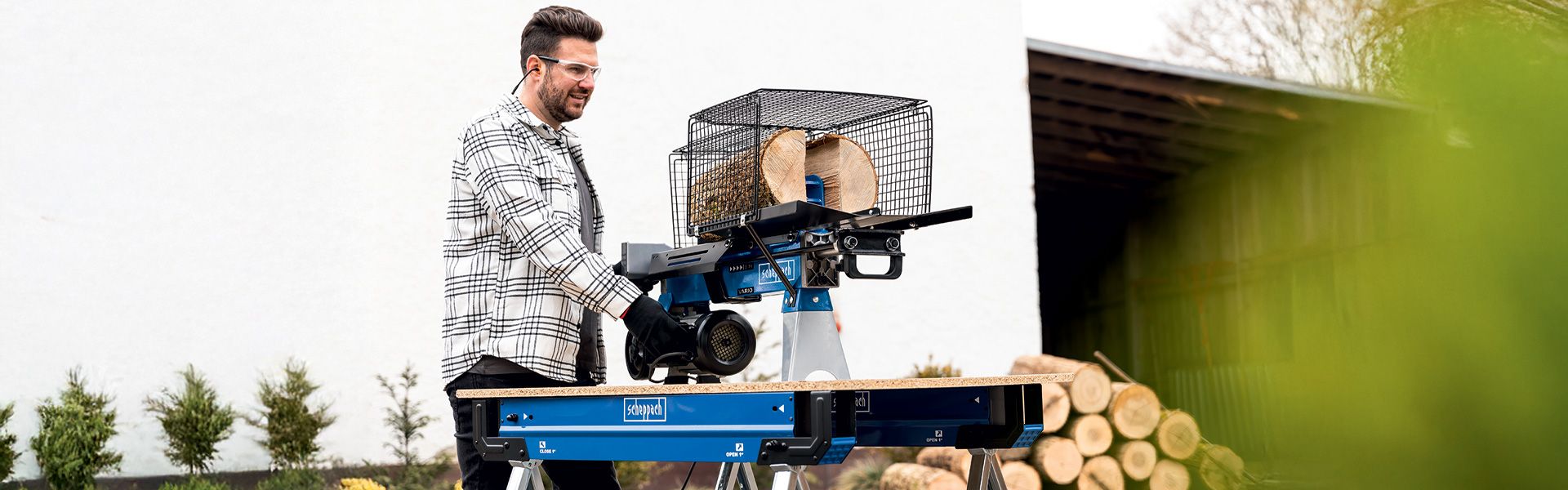 A man operates a Scheppach log splitter outdoors. Stacked firewood is visible in the background.