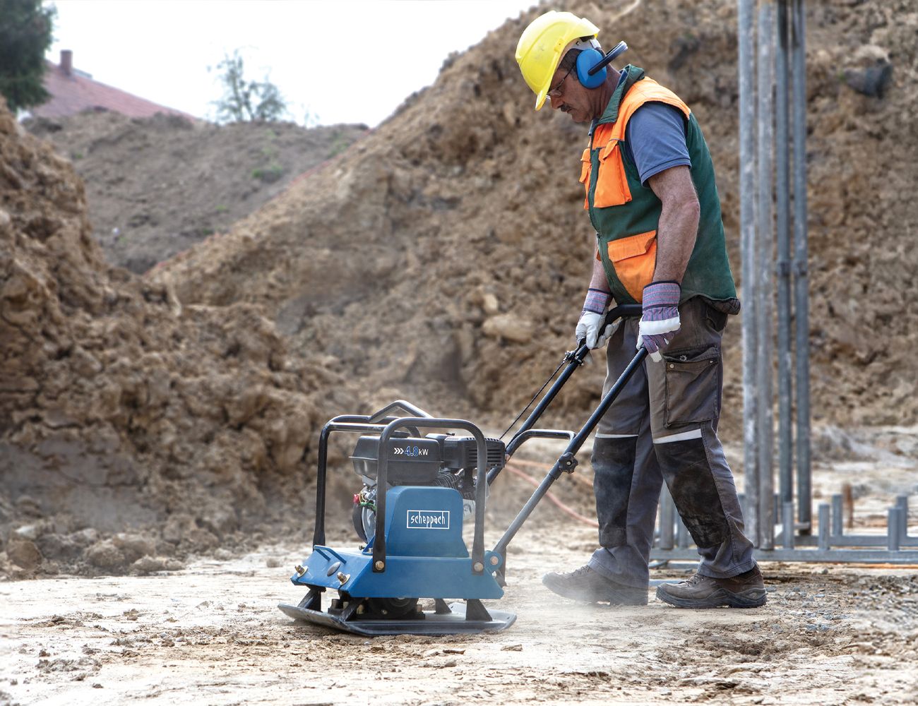 A worker in protective gear operates a Scheppach vibrating plate on a construction site. A mound of earth is visible in the background.