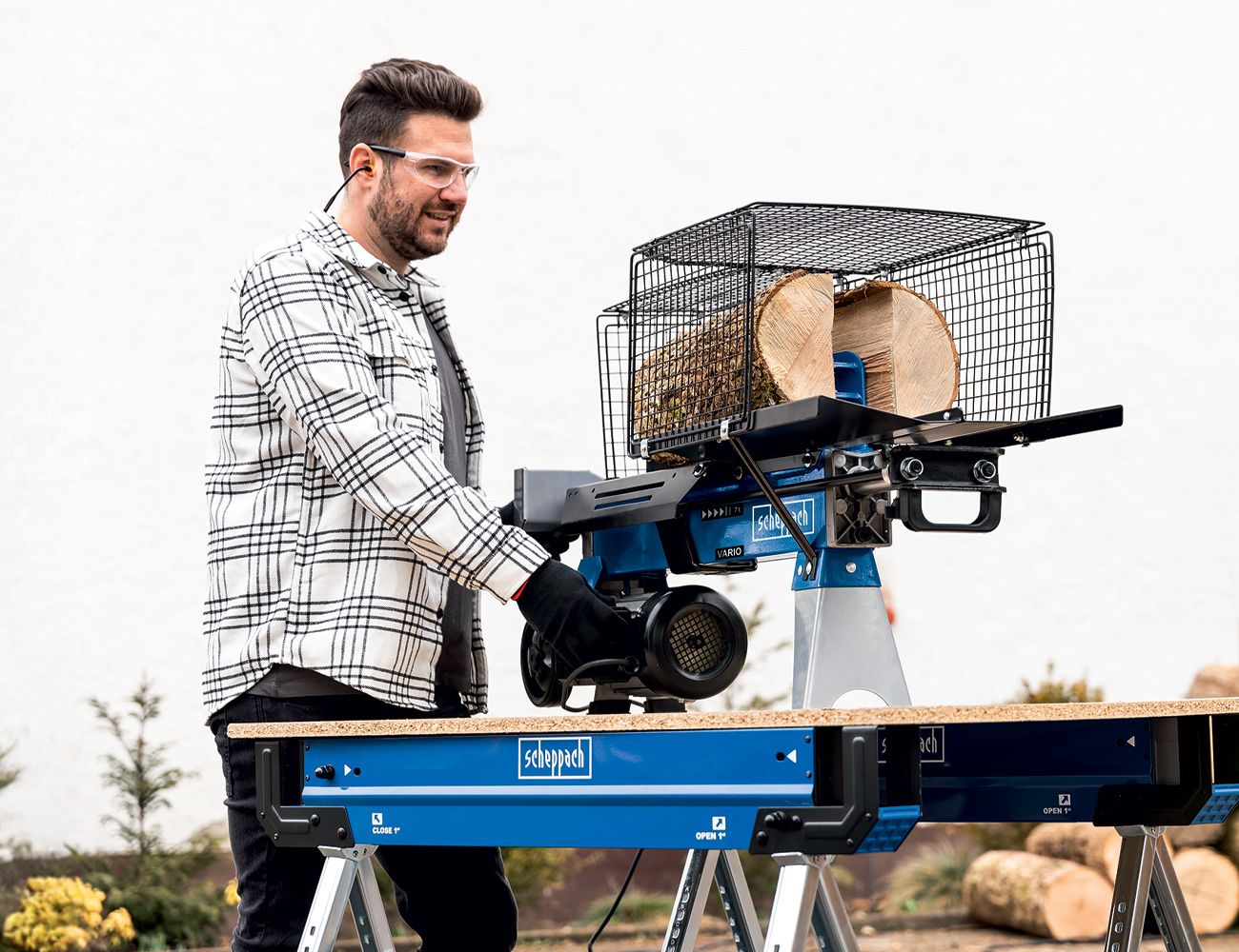 A man operates a log splitter by Scheppach. The machine is mounted on a sturdy table and has a basket for logs.