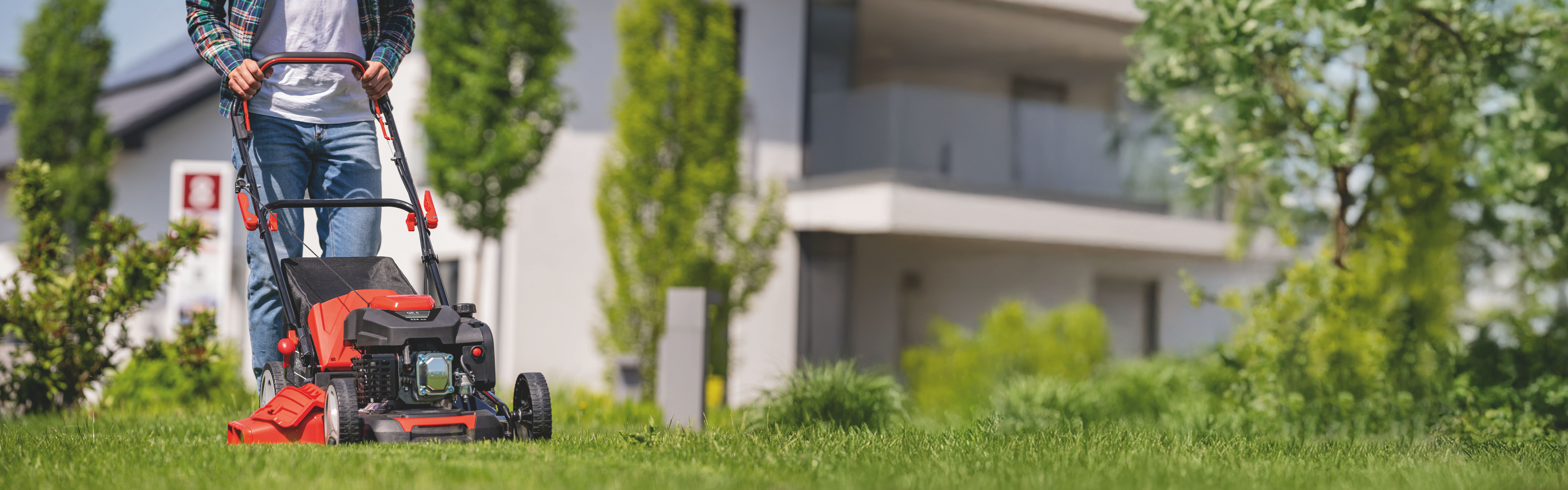 Ein Mann schiebt einen Rasenmäher auf einem grünen Rasen vor einem modernen Haus. Der Rasenmäher ist ein Scheppach-Produkt.
