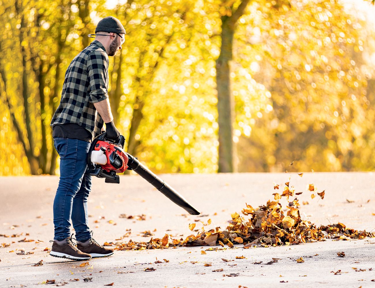 A man using a leaf blower in an autumn garden. Scheppach tools in action, surrounded by falling leaves. Autumnal atmosphere with golden foliage.